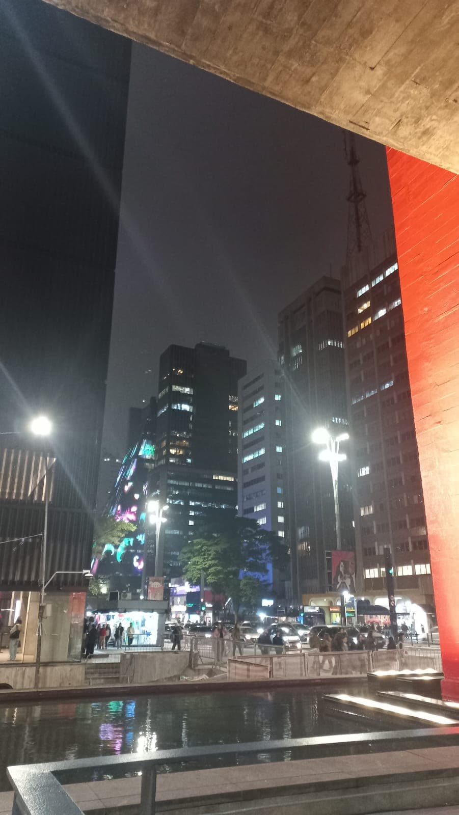 Cena urbana noturna com prédios iluminados, luzes e reflexos em espaço público.